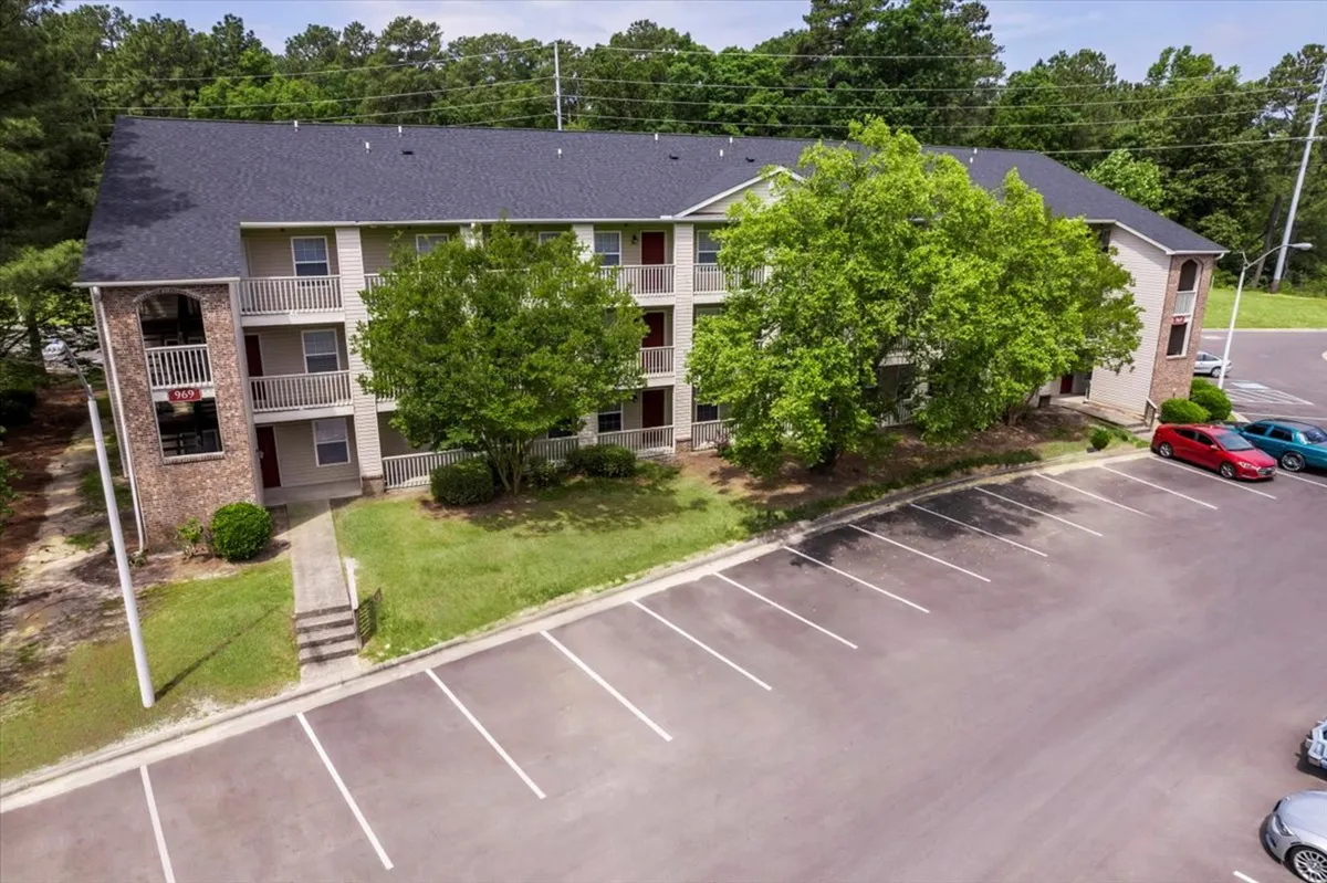 The photo shows a spacious, well-maintained parking lot surrounded by green trees and a modern apartment building. The area looks sunny and inviting, with a clear focus on easy access and the ability to come and go conveniently.