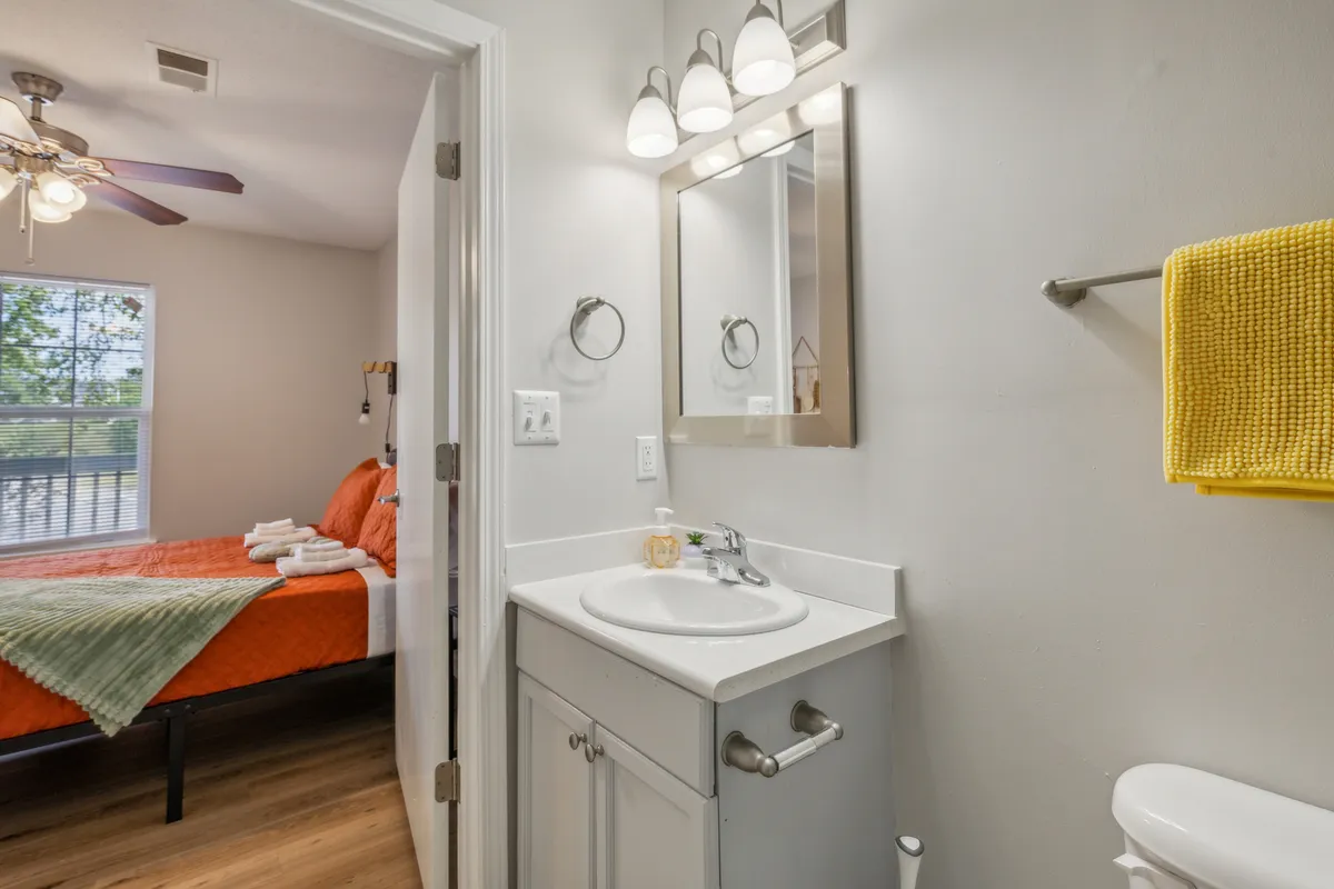 A cozy interior space with a vanity and sink, mirror, and fresh linens. An orange bedspread and a yellow towel add warmth and comfort, evoking the sense of thoughtful touches and attention to everyday needs.