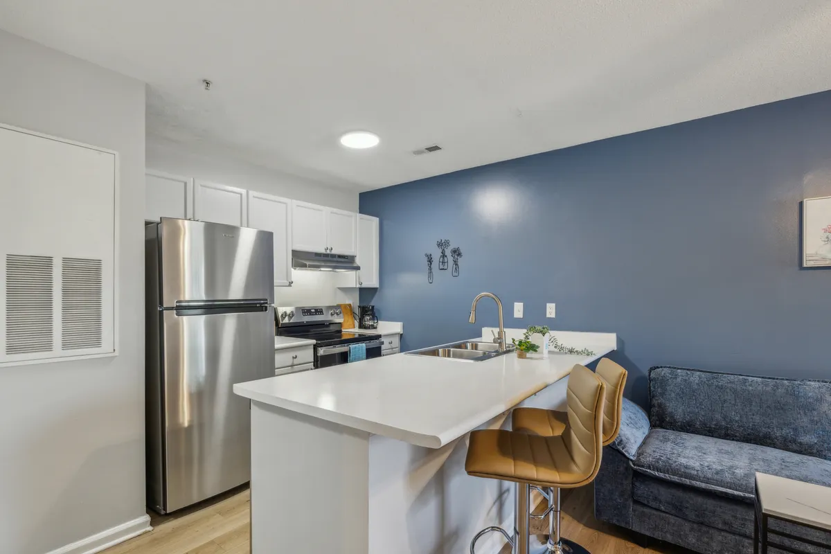 Another angle of the kitchen, highlighting the kitchen island surrounded by comfortable bar stools, stainless steel appliances, and the signature blue accent wall. The scene suggests a cozy spot for breakfast with family, sharing recipes, or entertaining guests in a homey environment.