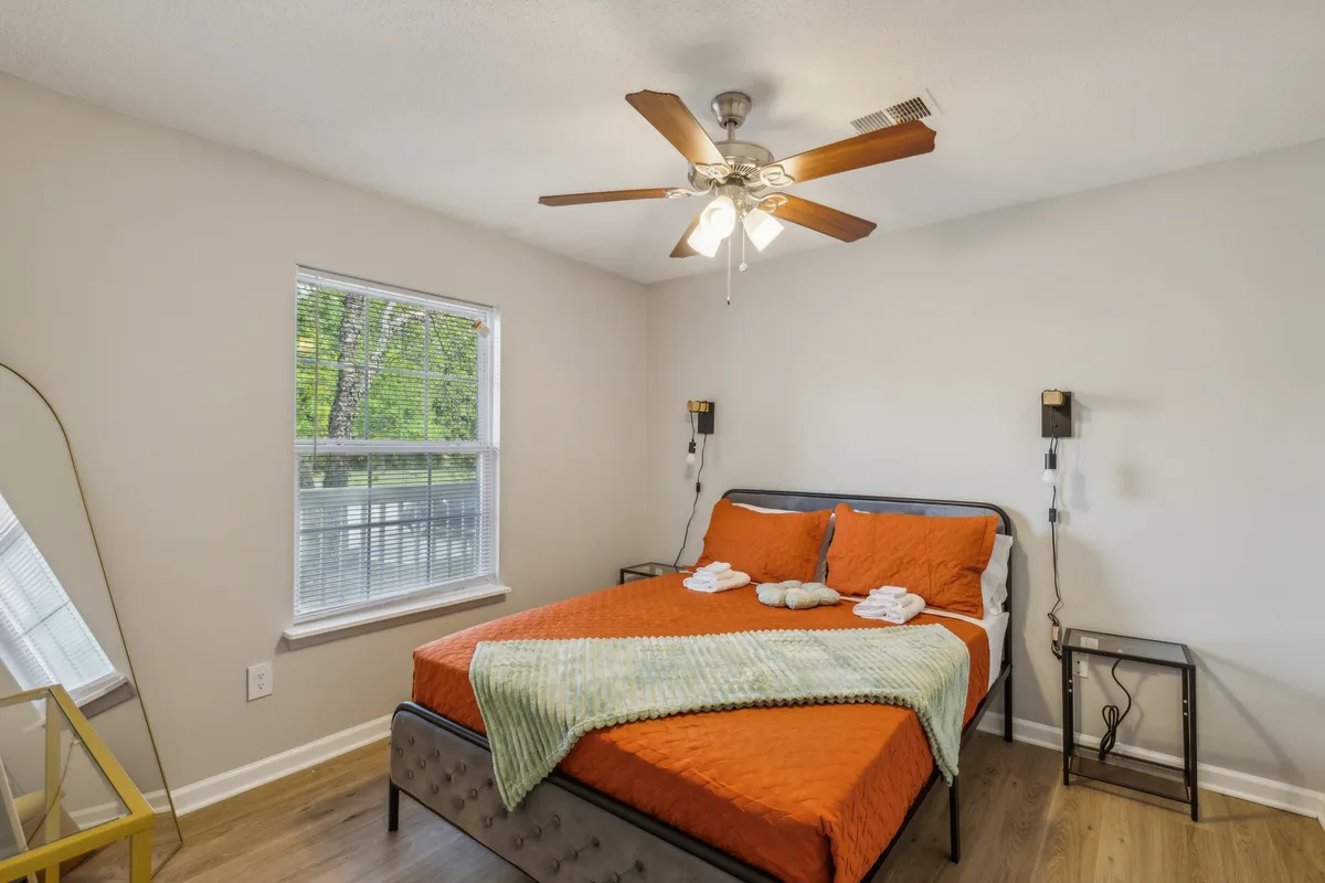 The image shows a welcoming bedroom featuring a plush bed with an inviting orange bedspread, flanked by modern nightstands topped with warm, glowing lamps. Soft natural light pours in through a large window, creating a bright and uplifting atmosphere ideal for relaxation or unwinding after a day of exploring Fayetteville.