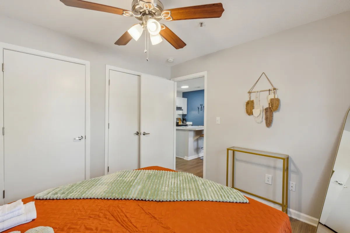 A cozy bedroom with an orange quilted bedspread, green textured throw blanket, ceiling fan with light fixture, and decorative wall hanging. The space feels warm and inviting, suggesting a restful environment ideal for sleep and relaxation.