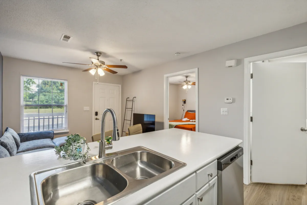 A view from the kitchen sink looking into the living room, showing the connection between cooking and social spaces. Natural light streams in from a window, and the layout combines kitchen activity with relaxation or entertaining in the adjoining area.