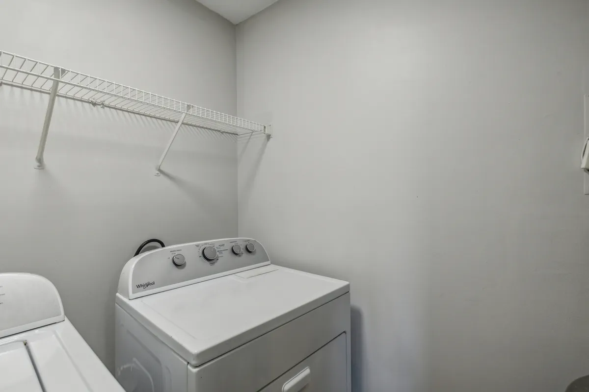 A dedicated laundry area featuring a modern washing machine, dryer, and a shelf above for storing laundry essentials. The scene is organized and practical, conveying the convenience of in-unit laundry and easy access to cleaning supplies.