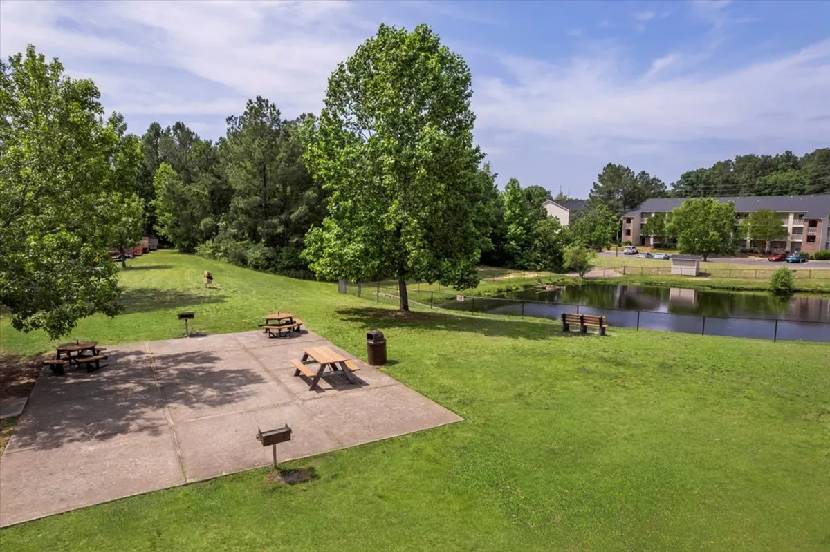 A peaceful outdoor scene featuring a pond surrounded by trees, benches, and a picnic area. Sunlight reflects off the water, creating a relaxing atmosphere ideal for quiet walks, outdoor dining, or sunset views.