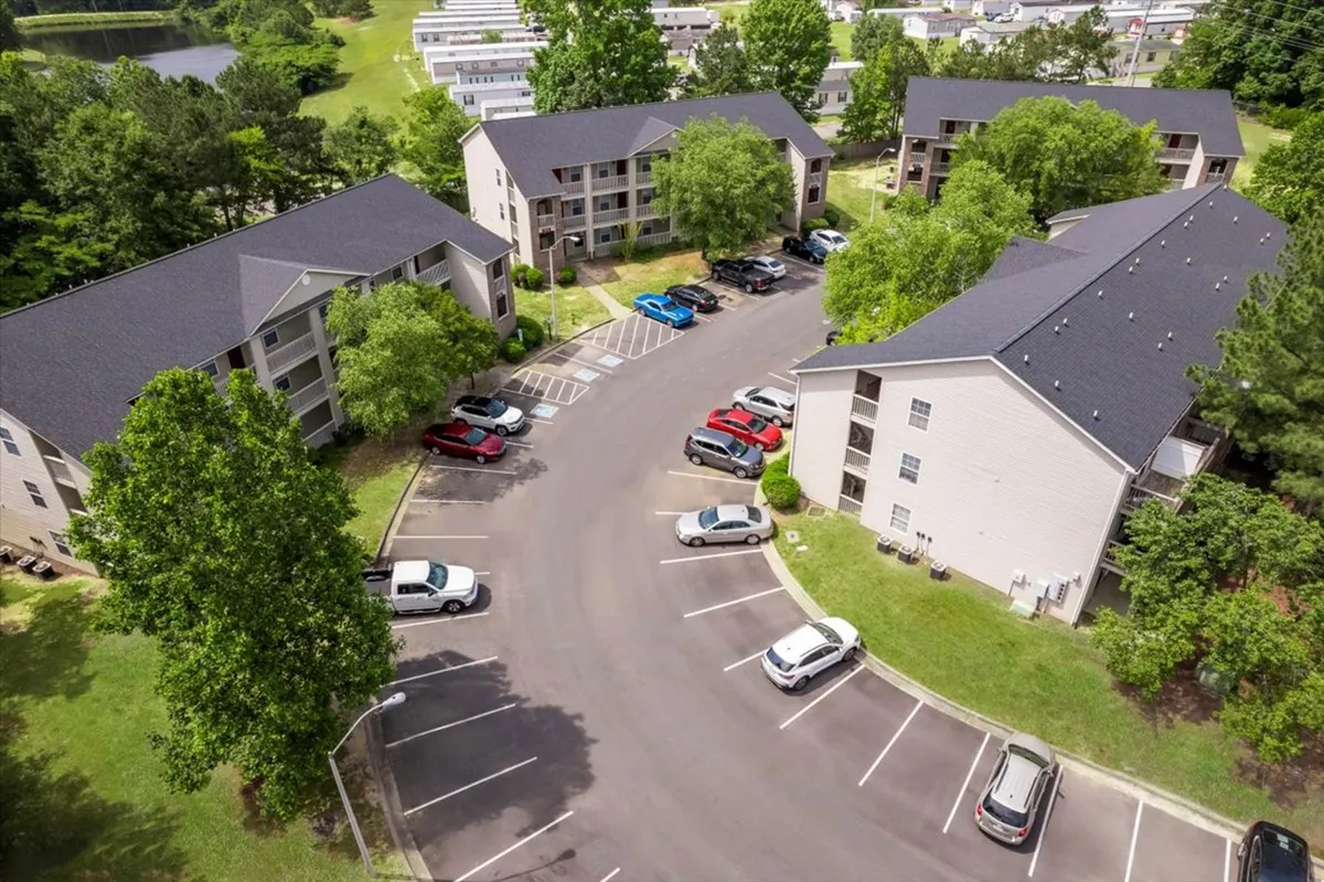 A wide view of the apartment complex, framed by mature green trees and a well-kept parking lot. Residents are seen socializing outdoors, with pathways and landscaping highlighting the connection between the apartments and the surrounding neighborhood.