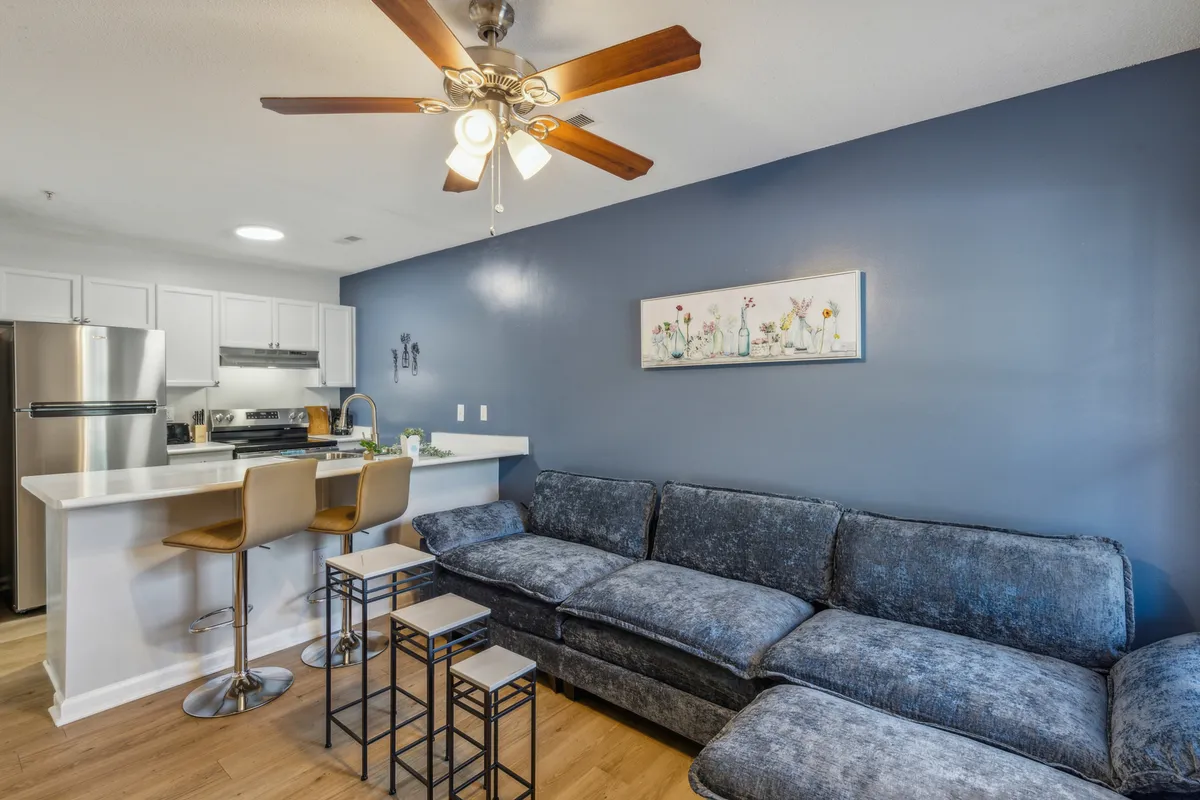 A cozy and inviting living area with a large sectional sofa, blue accent wall, kitchen bar area, and decorative artwork. The open layout and abundant natural light emphasize the spaciousness and homey feel.
