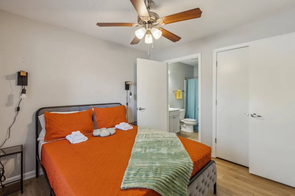 A view of the bedroom with orange bedding, ceiling fan, and the bathroom door visible, hinting at the proximity of the private bathroom. The room exudes comfort and convenience, with natural light filtering in.