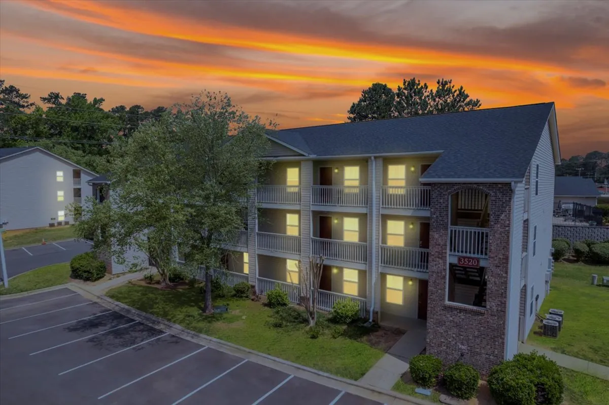 This exterior image captures the main building facade of the apartment complex at sunset, with warm light illuminating balconies and lush greenery all around. Residents are visible relaxing on their balconies and strolling in the peaceful grounds, creating a welcoming and tranquil atmosphere.
